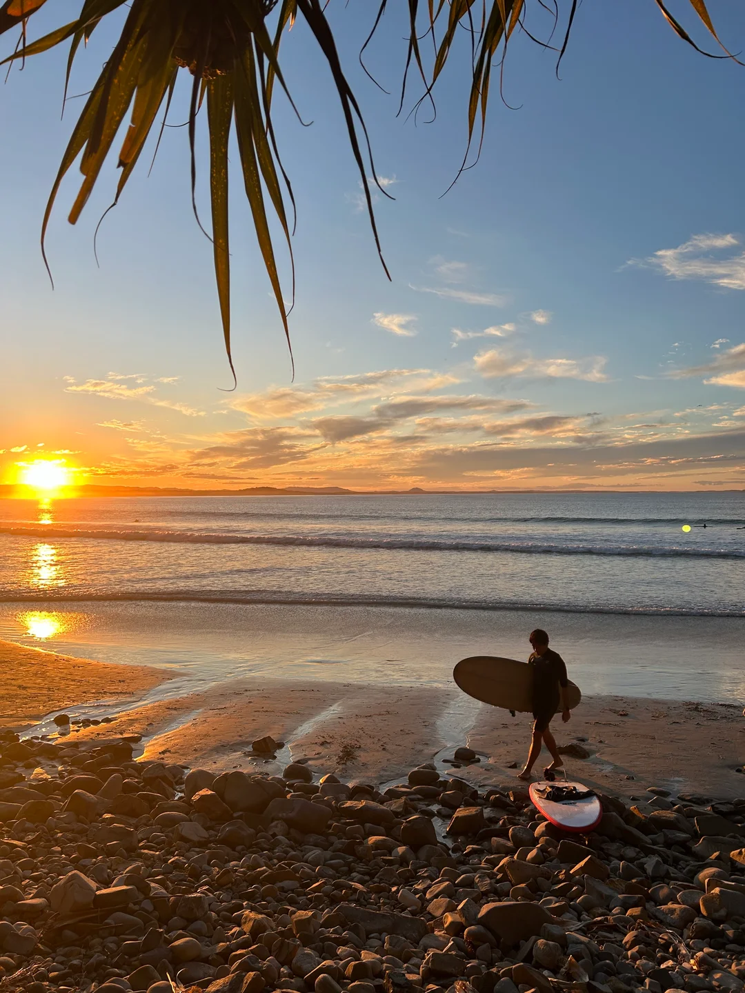 A surfer holding a board on a rocky beach, with a golden sunset over the waves.